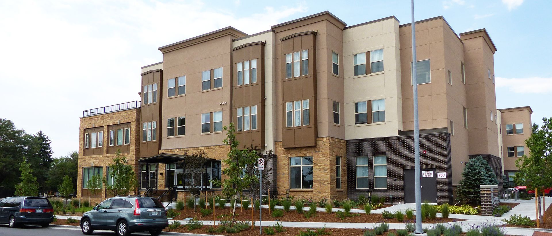 Tan apartment building with storefront-style windows, parked cars, and landscaping along a street