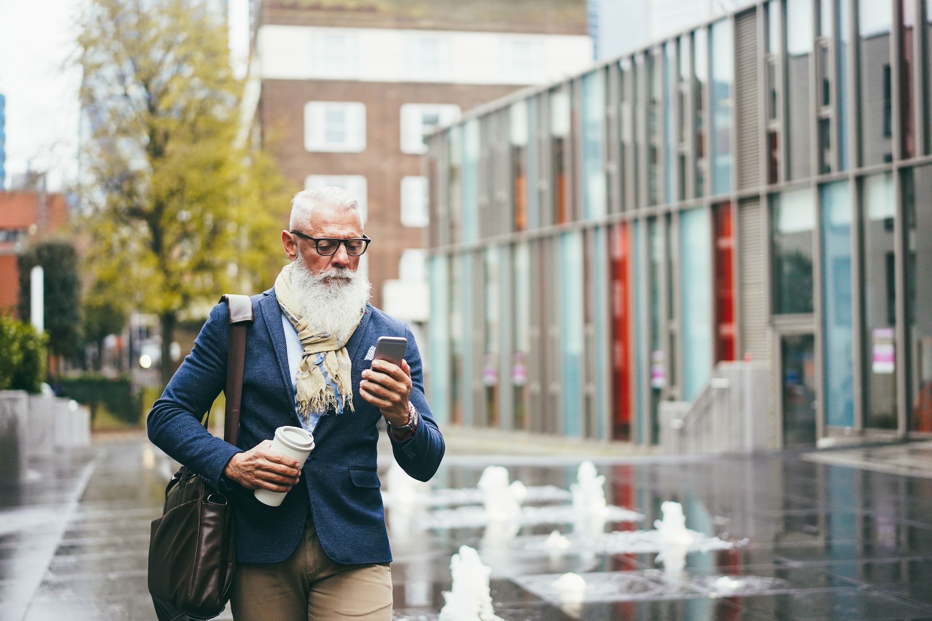 A man with a beard is walking down the street while looking at his cell phone.