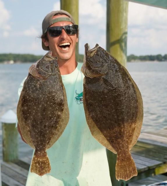 Man joyfully holds two large flounder fish on a dock by water. He wears sunglasses and a hat.