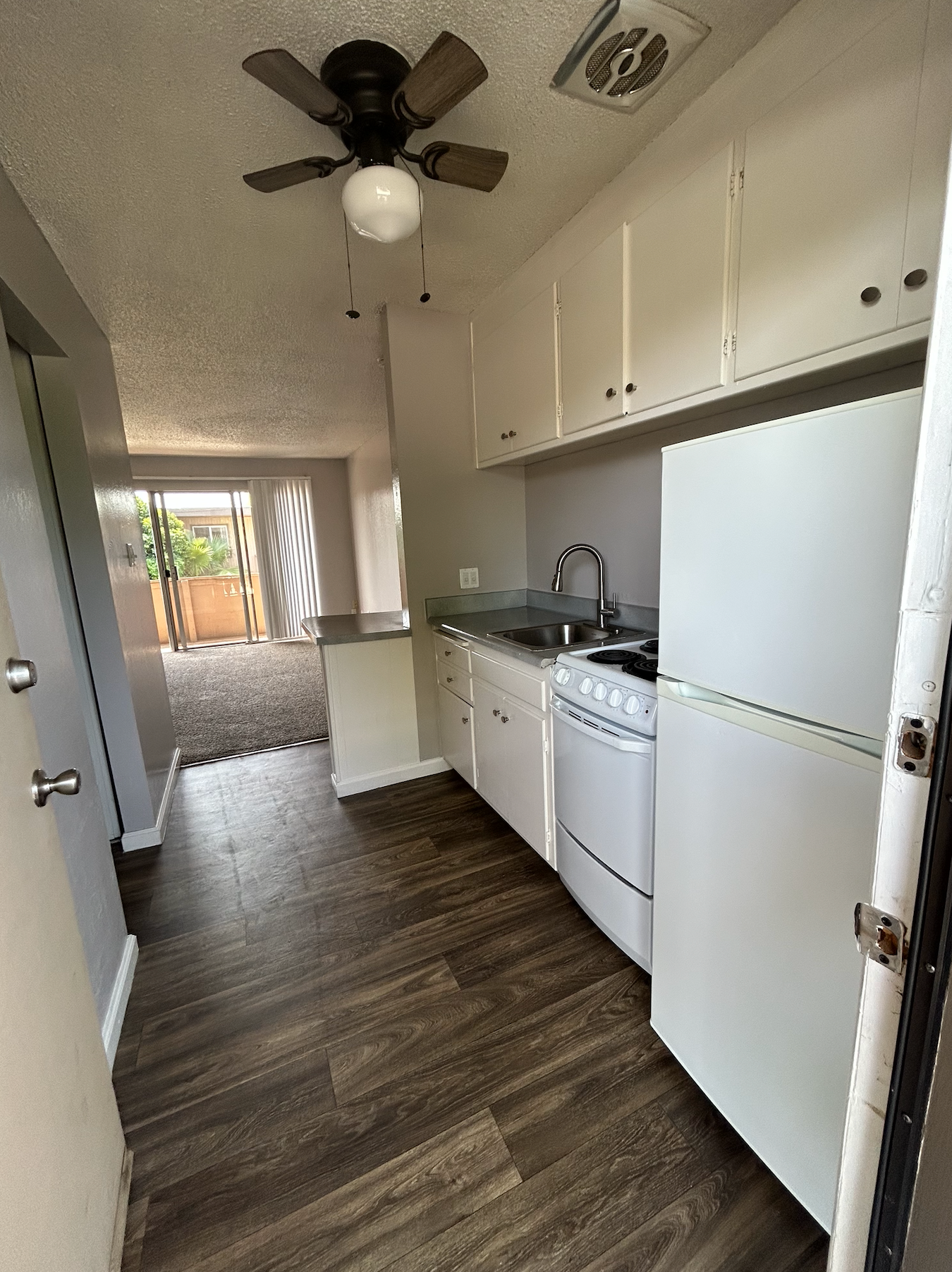 Narrow apartment kitchen with white cabinets, stove, refrigerator, ceiling fan, and wood-look flooring.