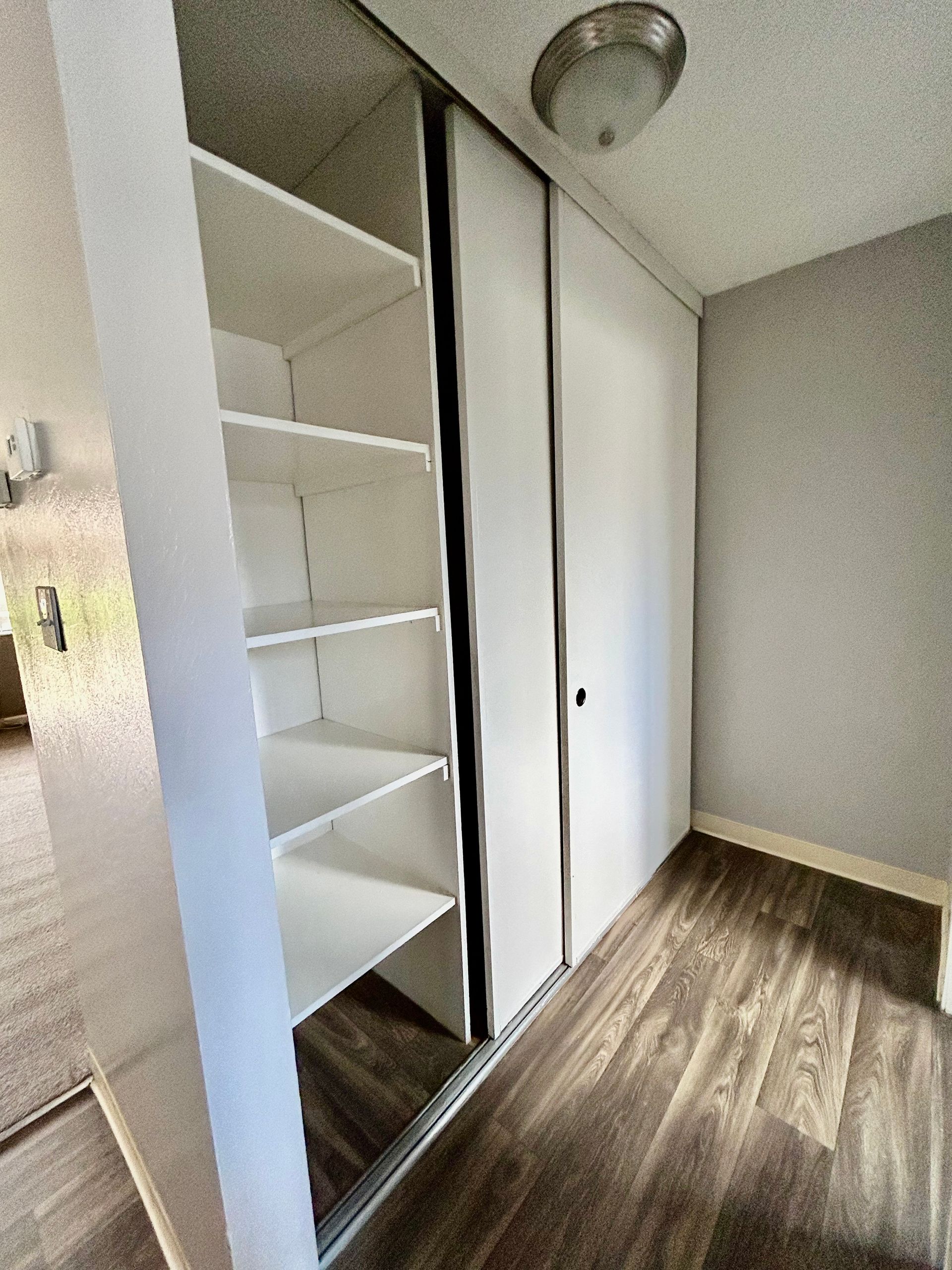 Small empty closet with white shelves, mirrored sliding door, and wood flooring.