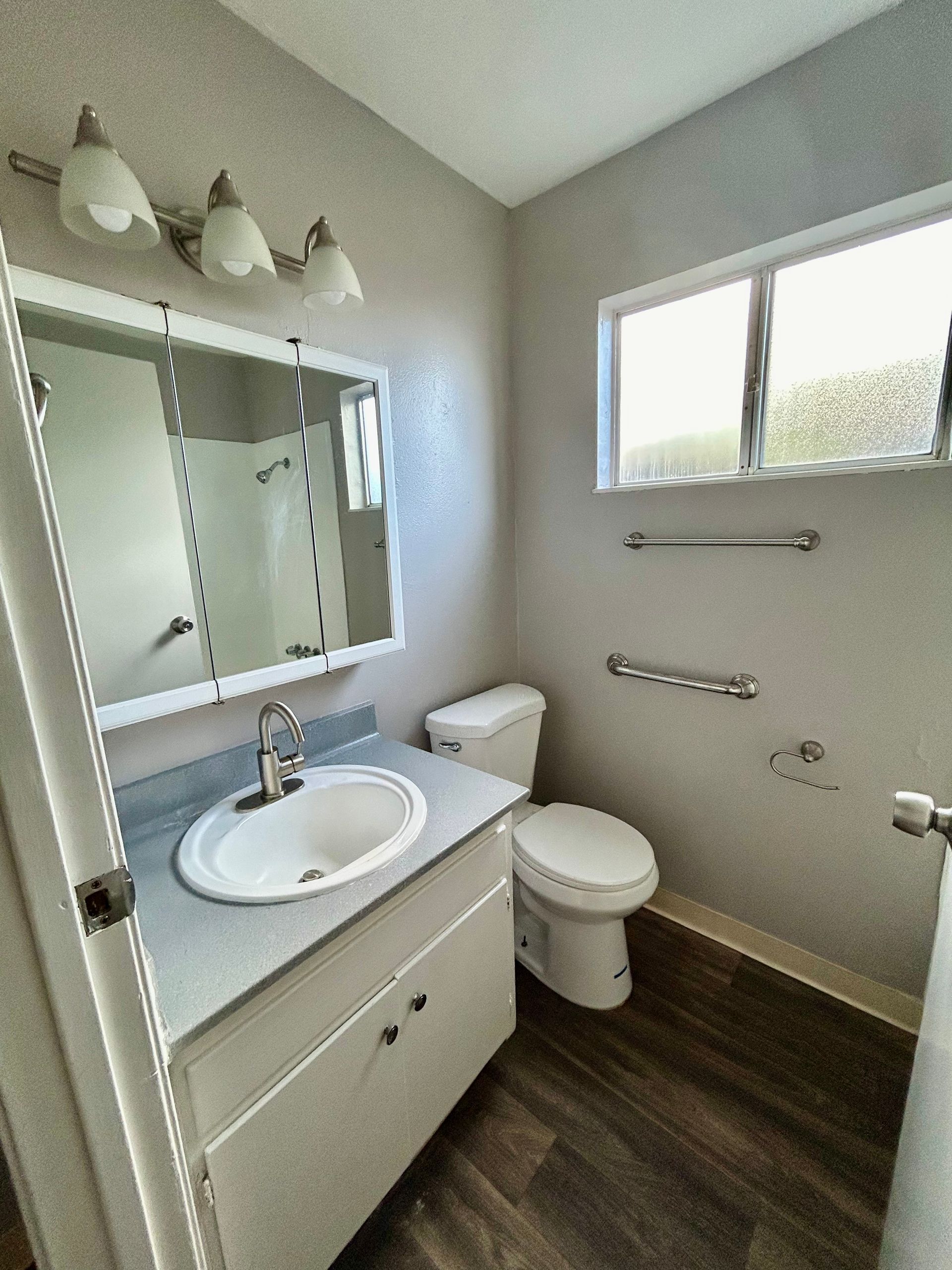 Small bathroom with white vanity, toilet, mirror cabinet, and frosted window on gray walls.