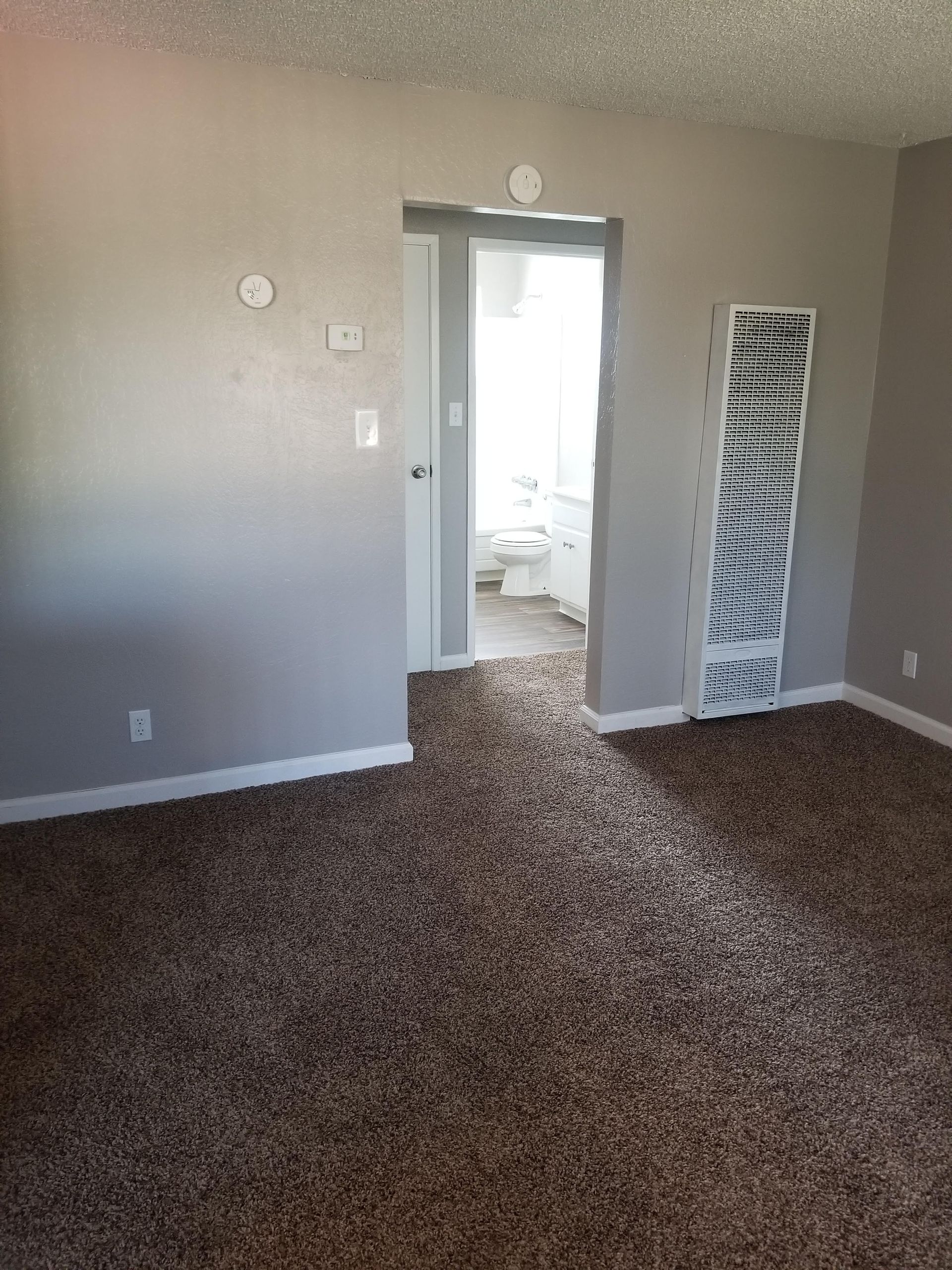 Empty beige carpeted room with open doorway to a bathroom and mirrored closet doors.