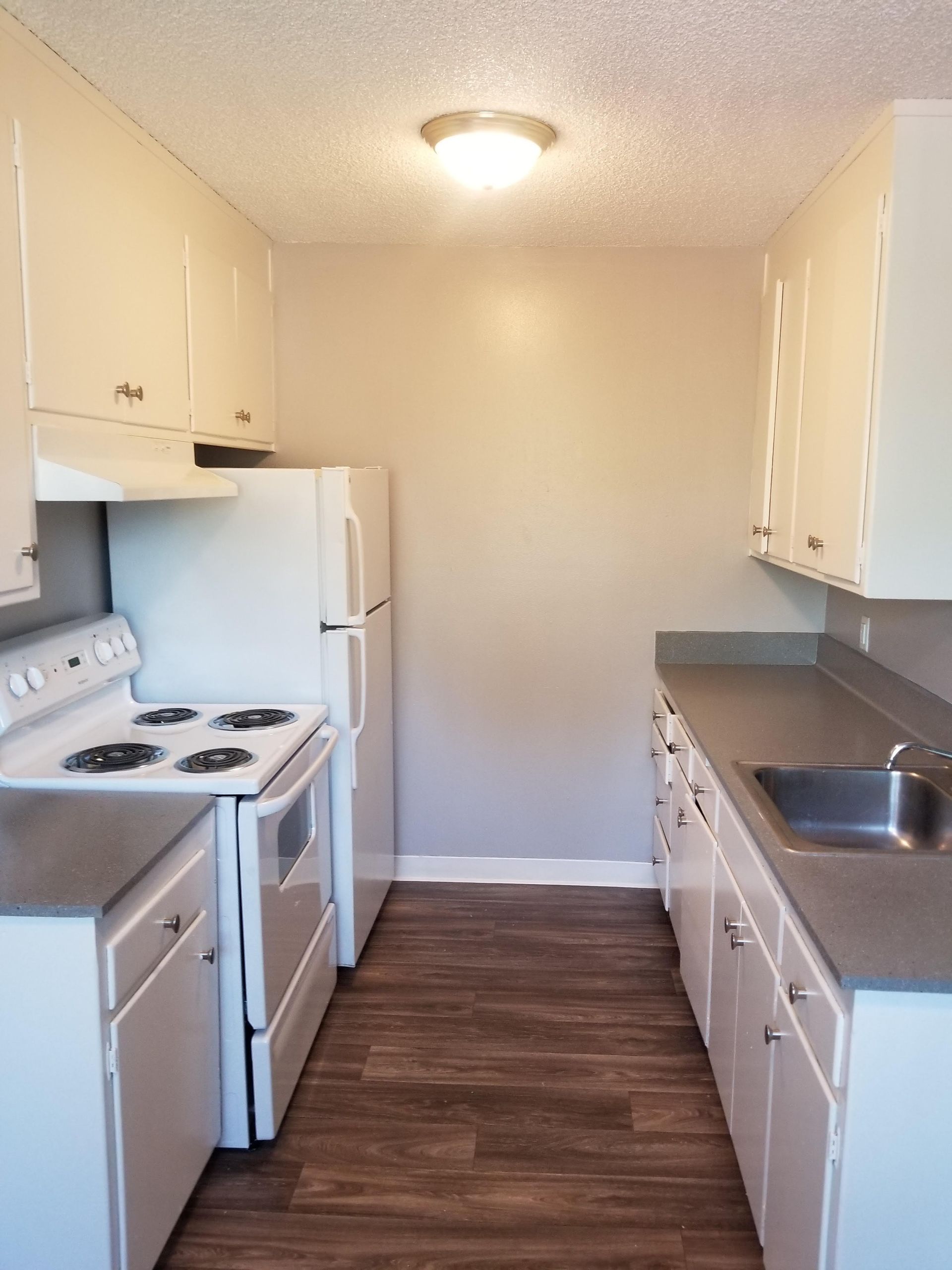 Small galley kitchen with white cabinets, stove, refrigerator, sink, and dark wood flooring.
