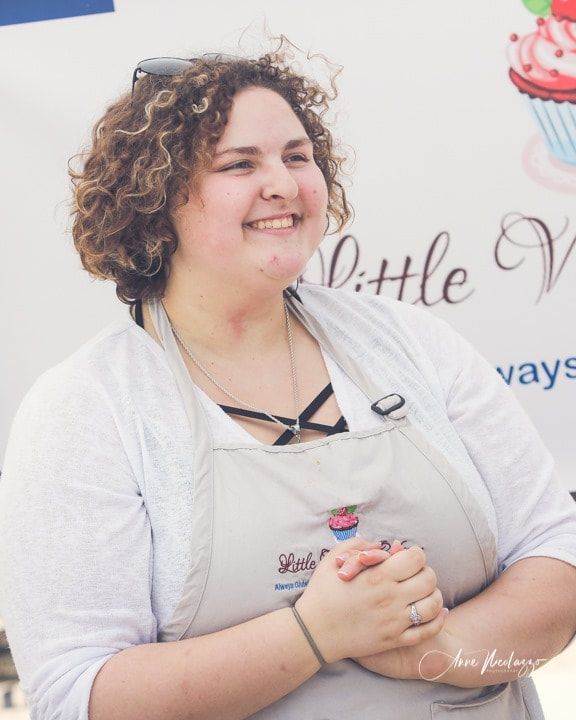 A woman wearing an apron is smiling in front of a sign