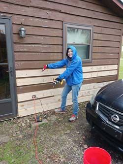 Person in a blue jacket holding a hose beside a house, with a red bucket and black car nearby.