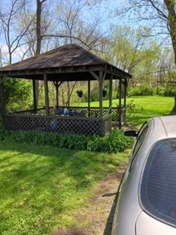 Wooden gazebo with a dark roof in a grassy yard beside a silver car on a sunny day