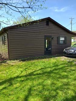 Brown single-story house with a black door and car parked beside it on a sunny lawn.