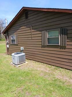Brown house exterior with window shutters and a central air unit on a grassy yard