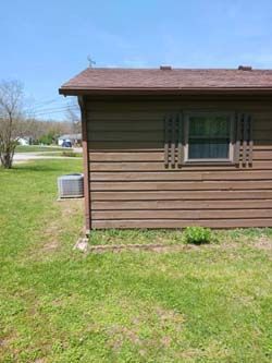 Side view of a brown house with a window air conditioner, set beside a grassy yard under a blue sky