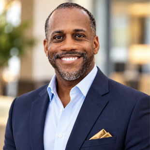 Smiling man in a navy suit with a light blue shirt and gold pocket square, standing outdoors.