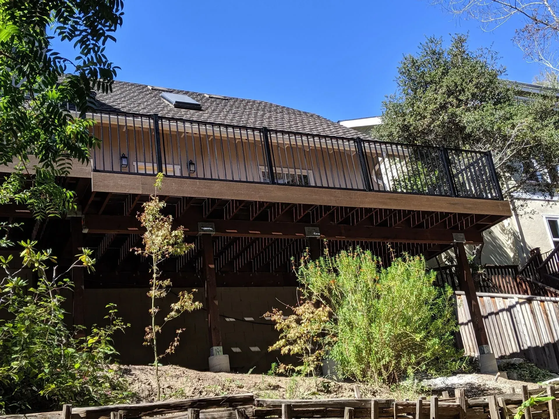 Two-story house with a weathered roof and balcony, partly hidden by lush green trees under a clear blue sky