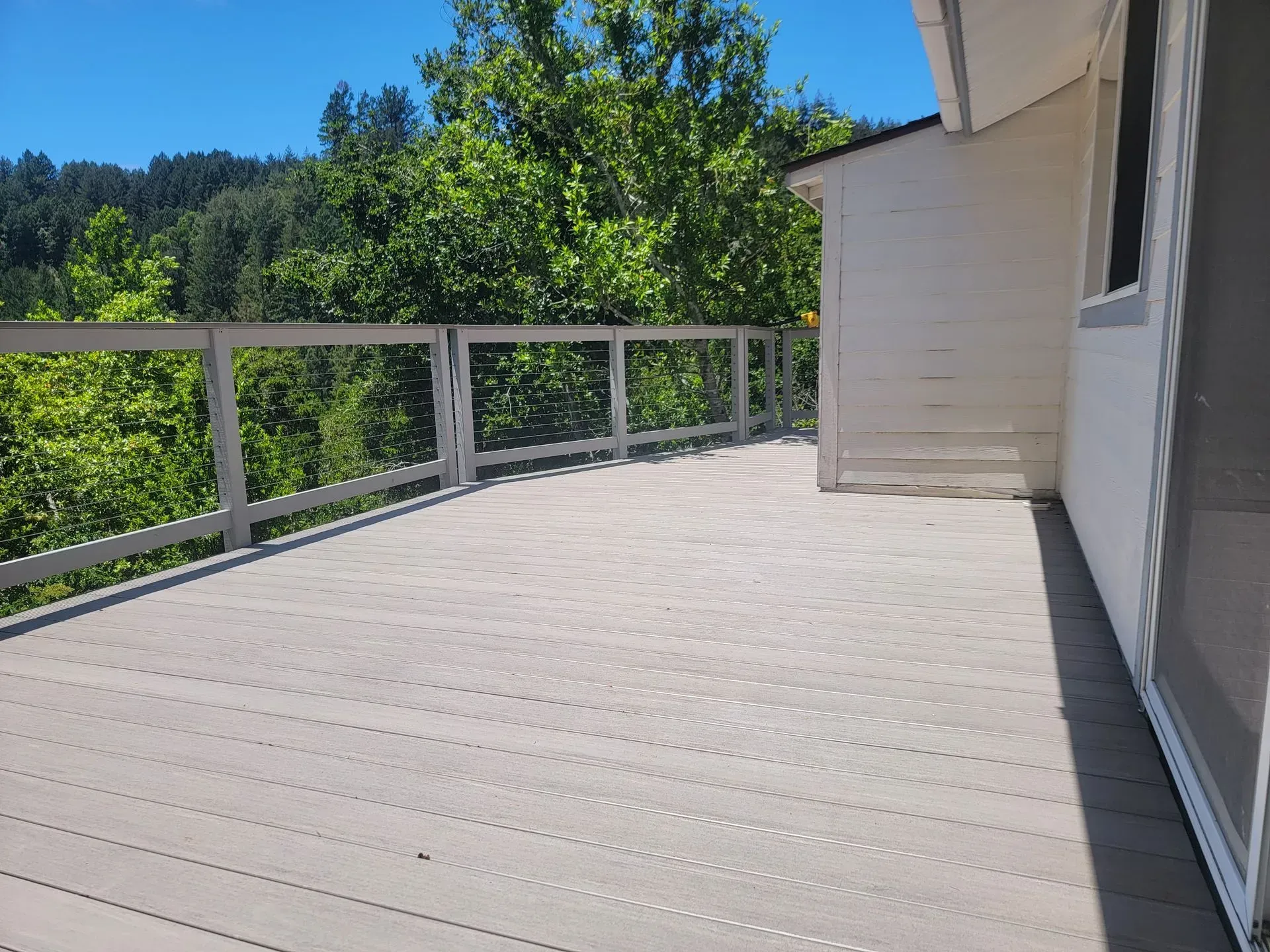 Sunlit wooden deck with white railing beside a house, overlooking green trees and hills