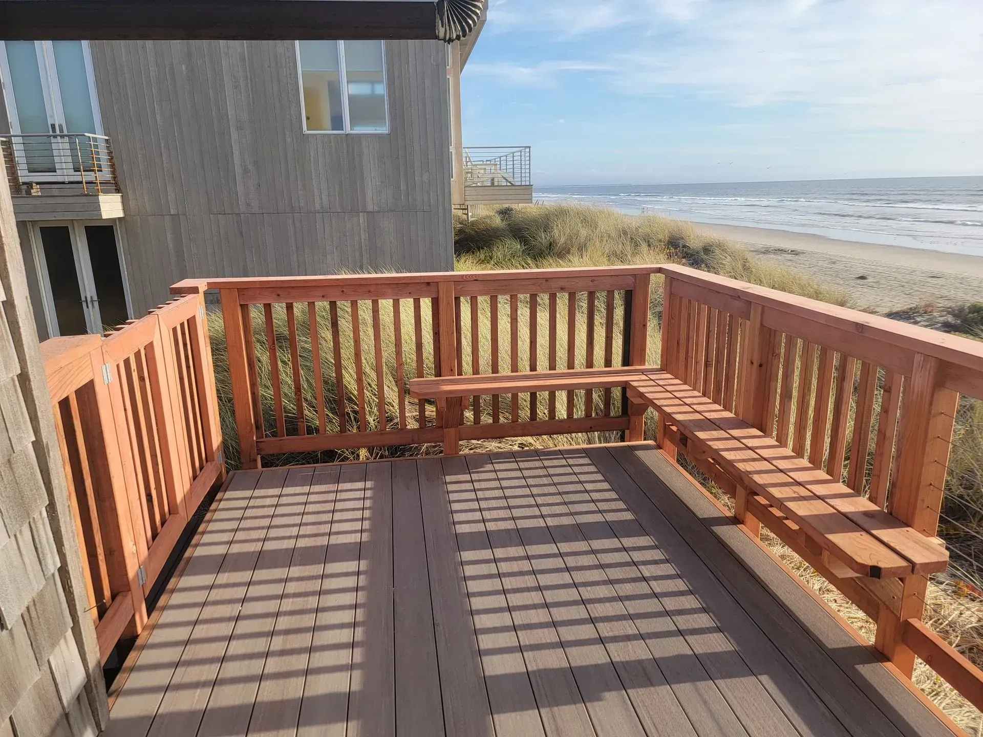 Wooden ocean-view deck beside a beach house, with benches and railings in sunlight.