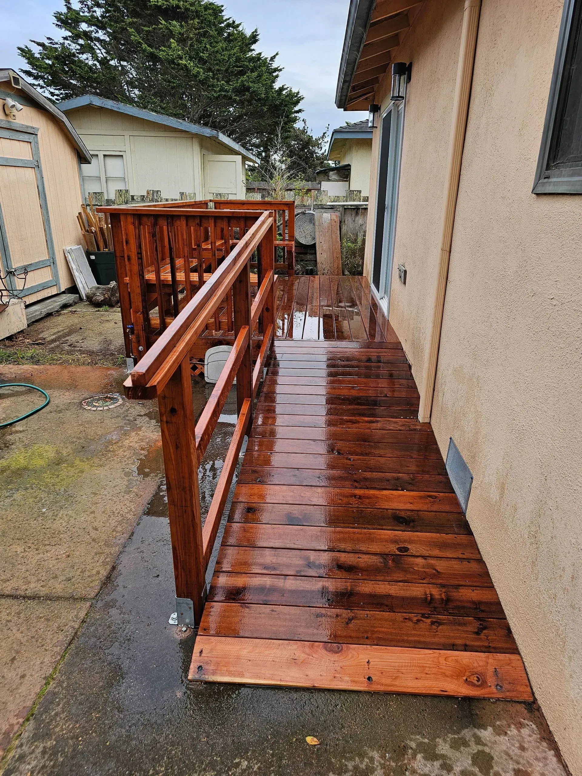 Wet wooden ramp beside a house, with railings and a narrow walkway in a backyard.