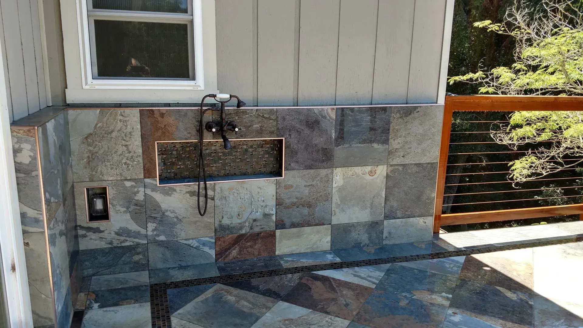 Outdoor wall-mounted faucet and stone sink beneath a window, with a tiled patio and wood railing.