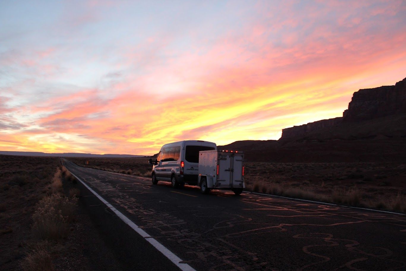 A van and trailer are parked on the side of a road at sunset.