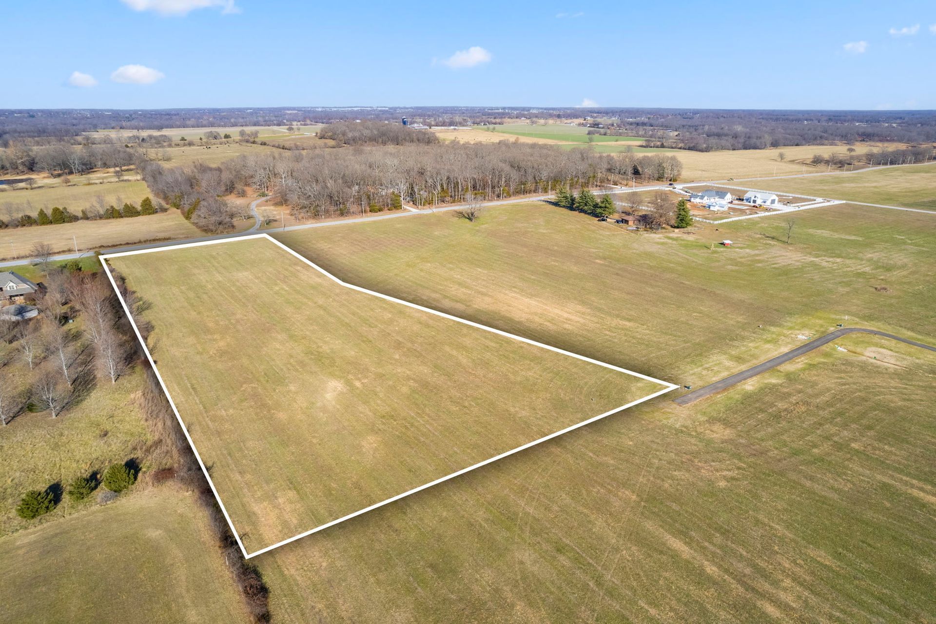 Aerial view of a large fenced grassy field beside trees and a few buildings under a blue sky