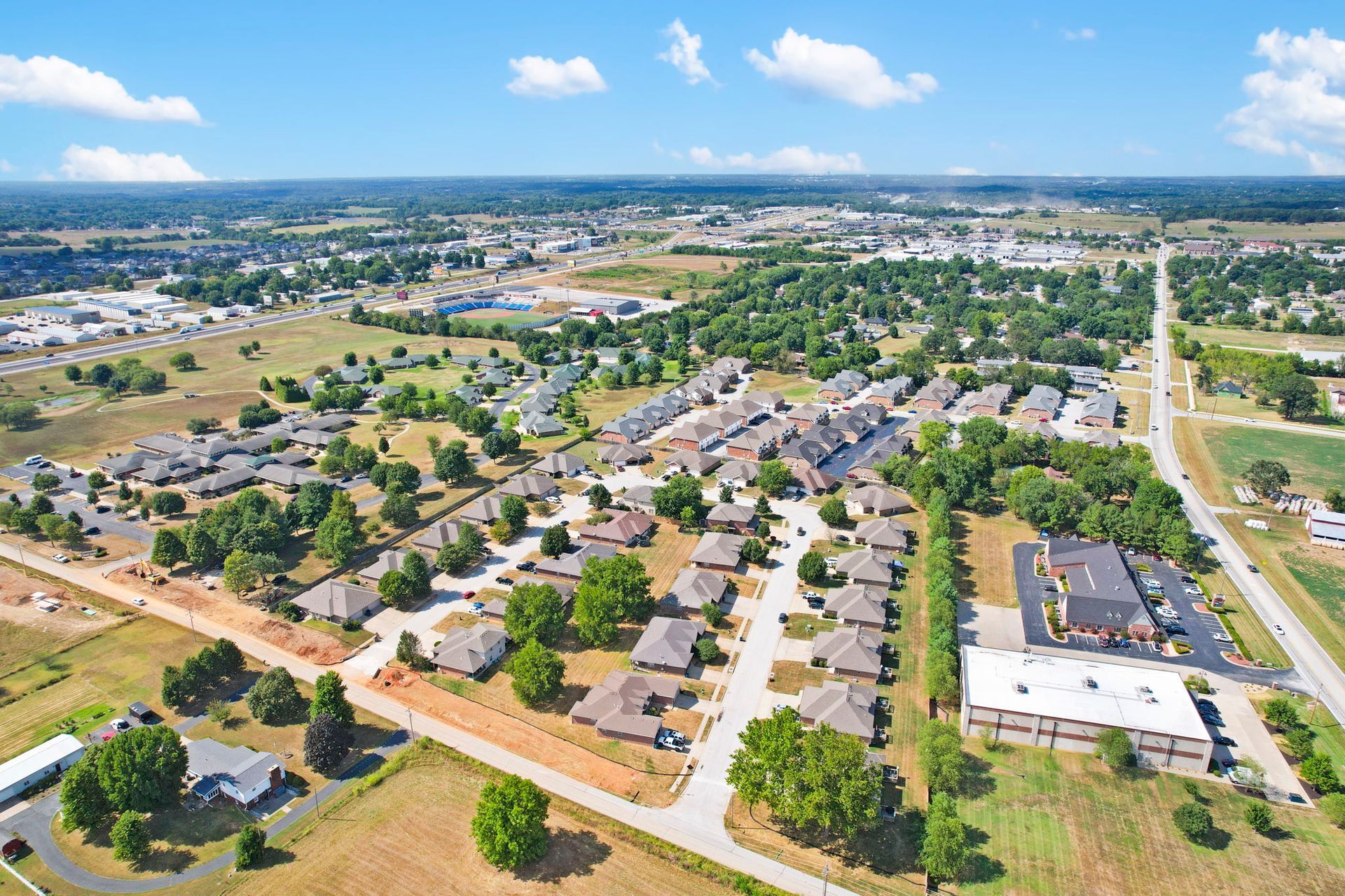Aerial view of a suburban neighborhood with houses, roads, trees, and a large building under a blue sky