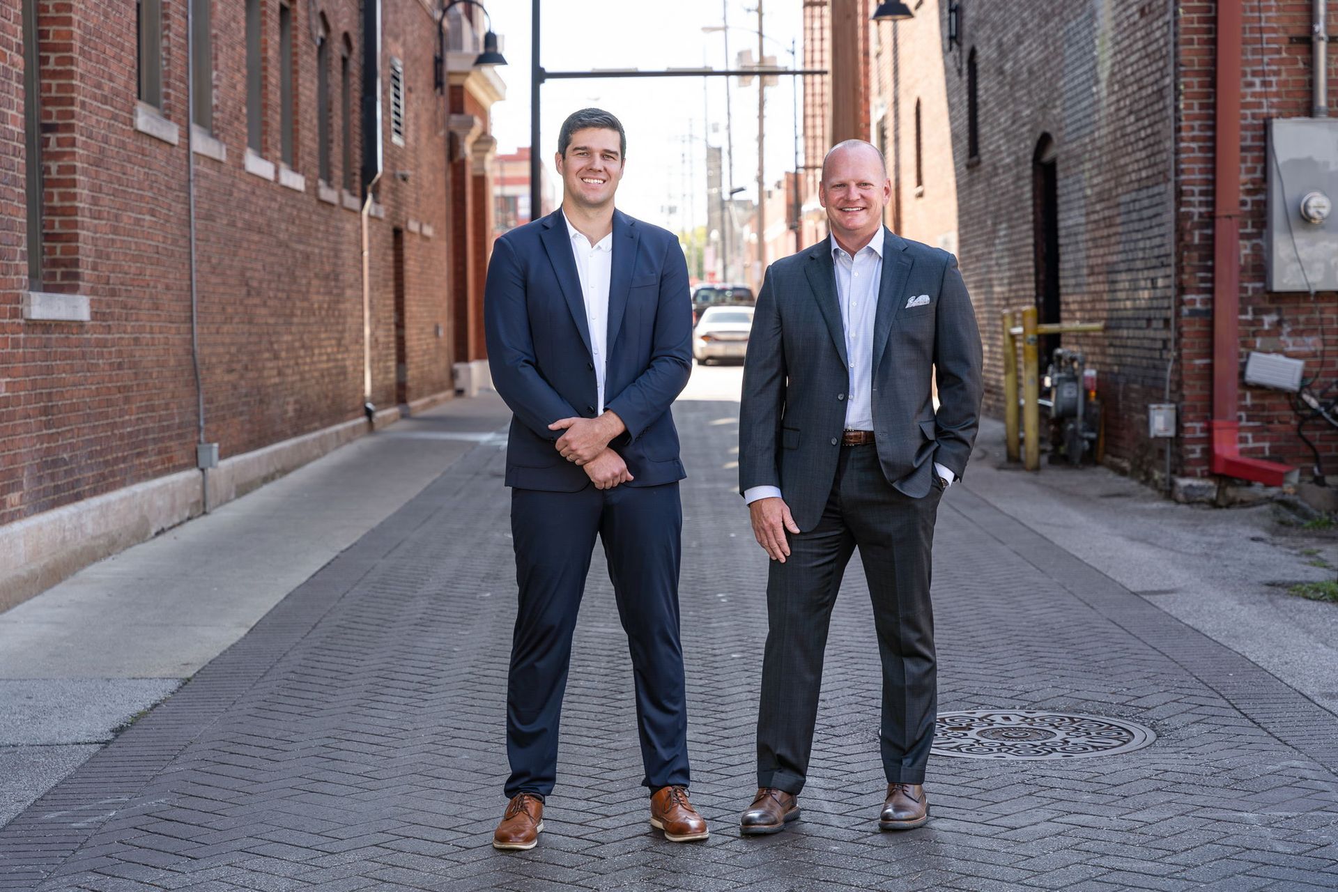 Two men in suits standing on a cobblestone alley between brick buildings