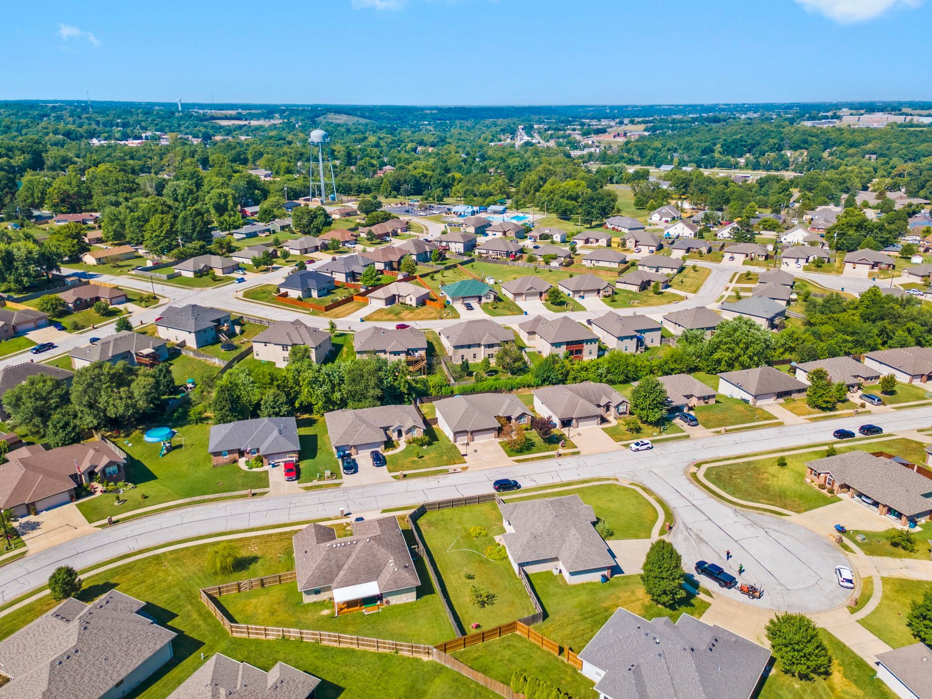 Aerial view of a suburban neighborhood with houses, streets, and green yards under a blue sky.