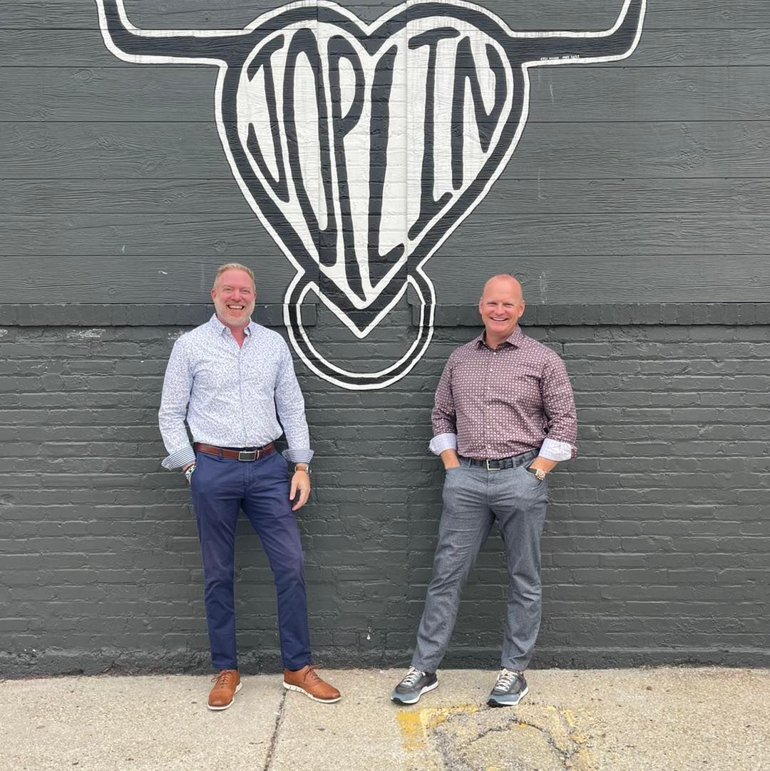 Two men standing in front of a black brick wall with a large white heart mural
