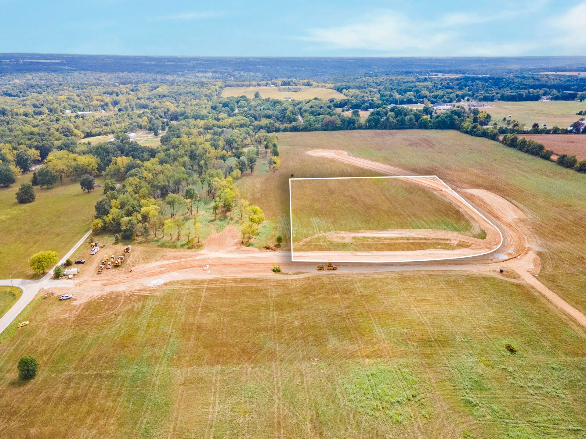 Aerial view of a dirt road curving through a grassy rural field with trees and farmland beyond