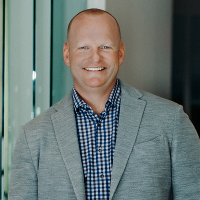 Smiling man in a gray blazer and blue checkered shirt standing indoors against a glass wall