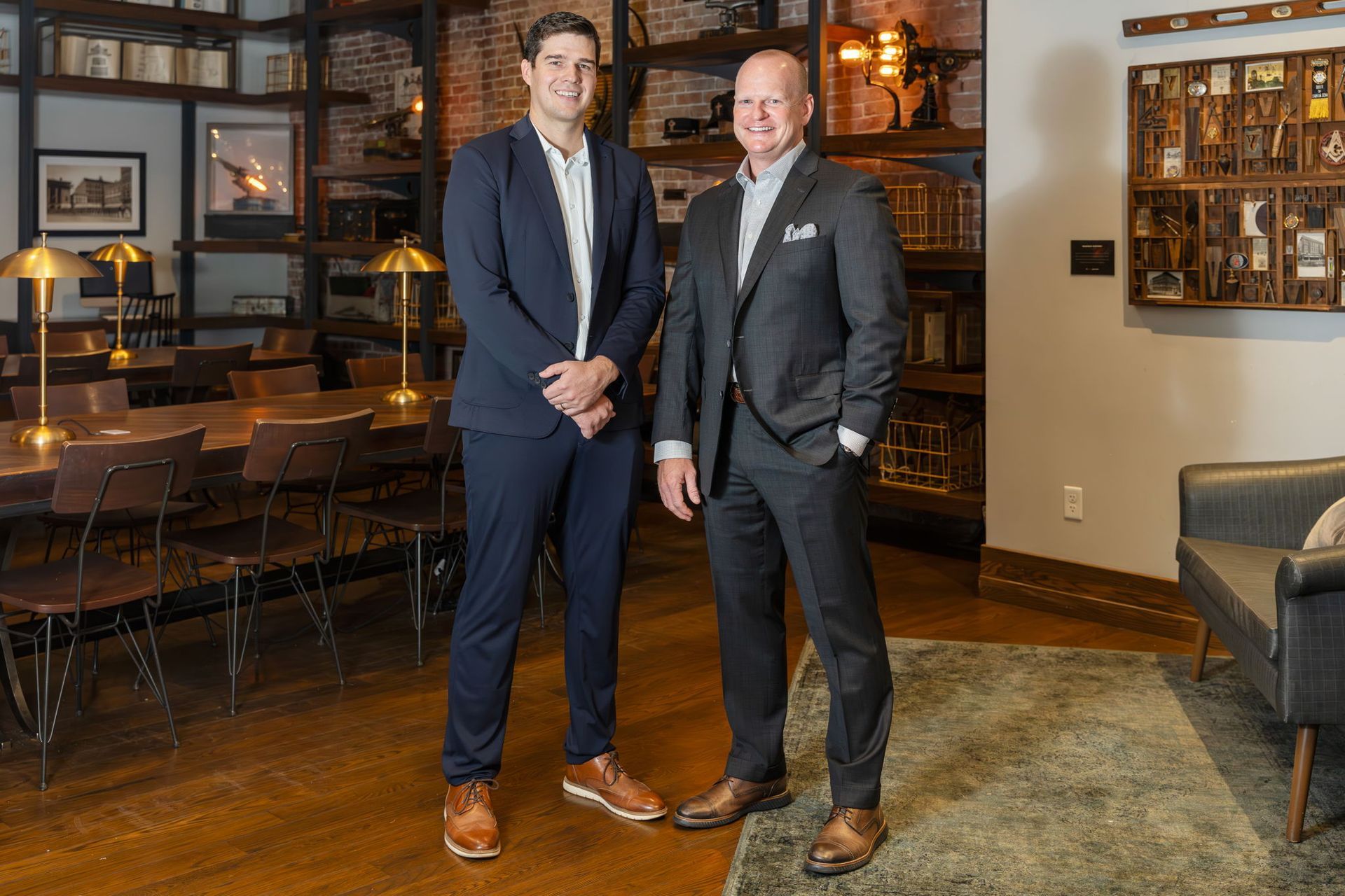 Two men in suits standing in a warmly lit lounge with framed art and leather seating.