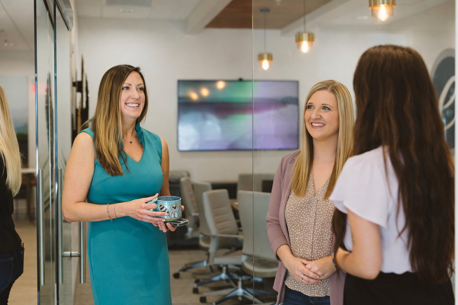 Three women chatting in a bright office meeting room, with chairs and a wall-mounted screen in the background