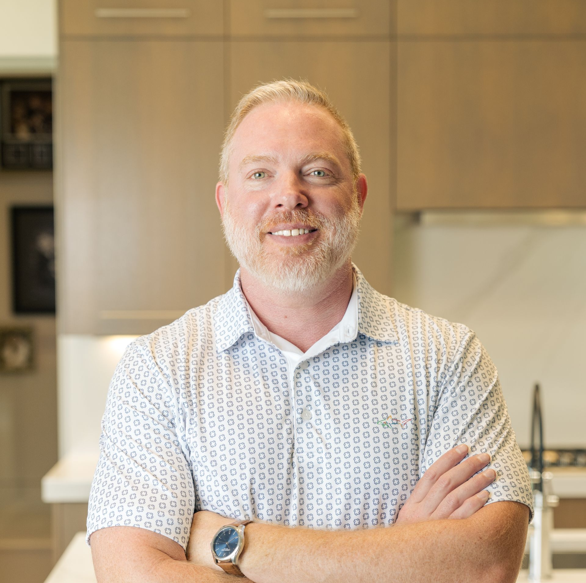 Man smiling with arms crossed in a bright kitchen, wearing a patterned shirt and watch.