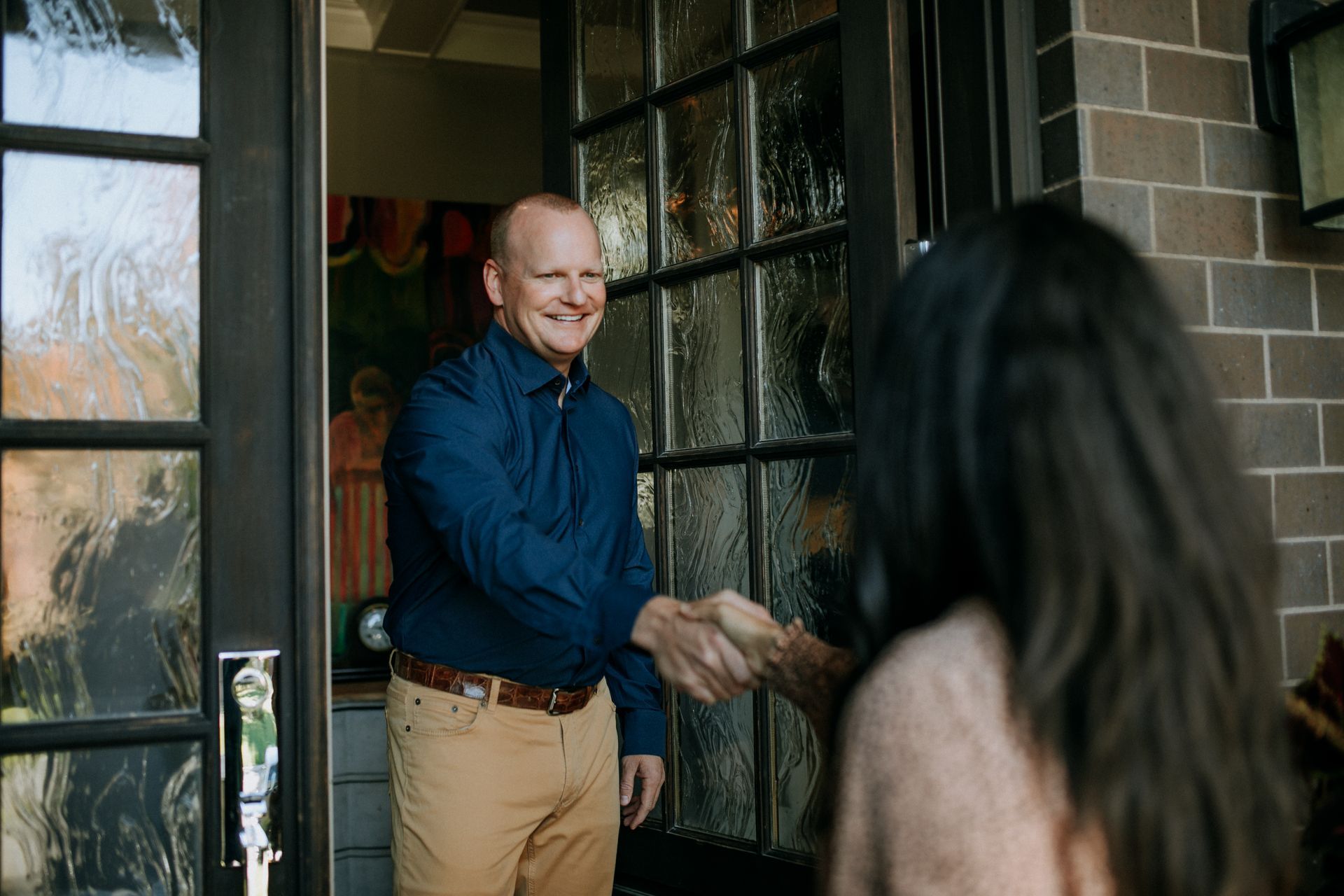 A man in a blue shirt shakes hands at a front doorway with a woman entering the house.