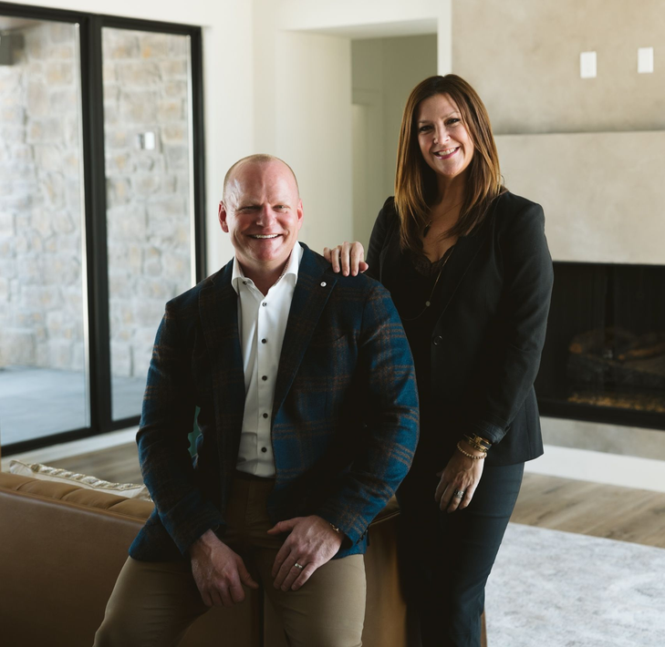 Two people in business attire smiling in a modern living room near a fireplace and large windows