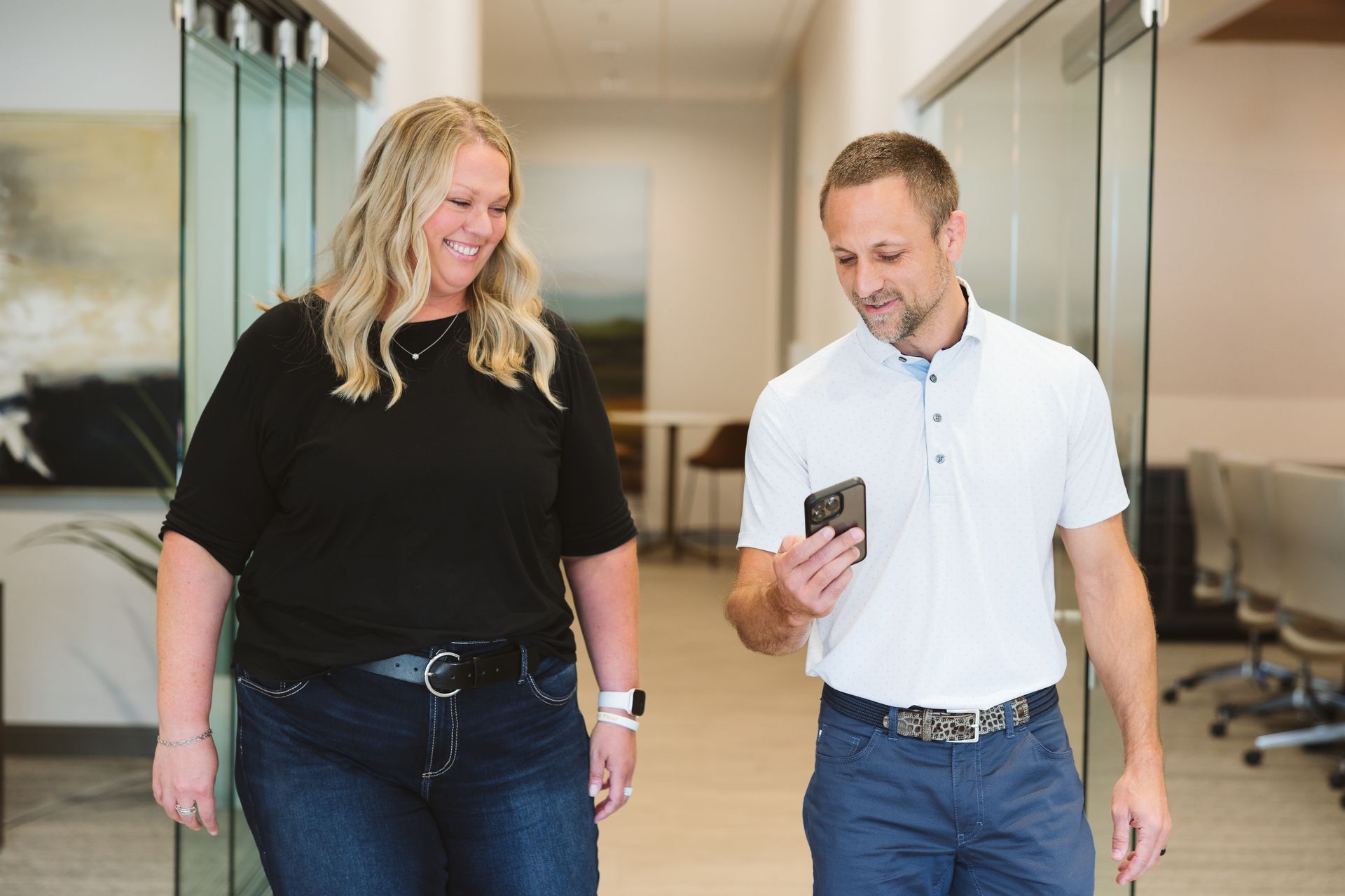 Two people walking down a hallway, smiling and looking at a phone together.