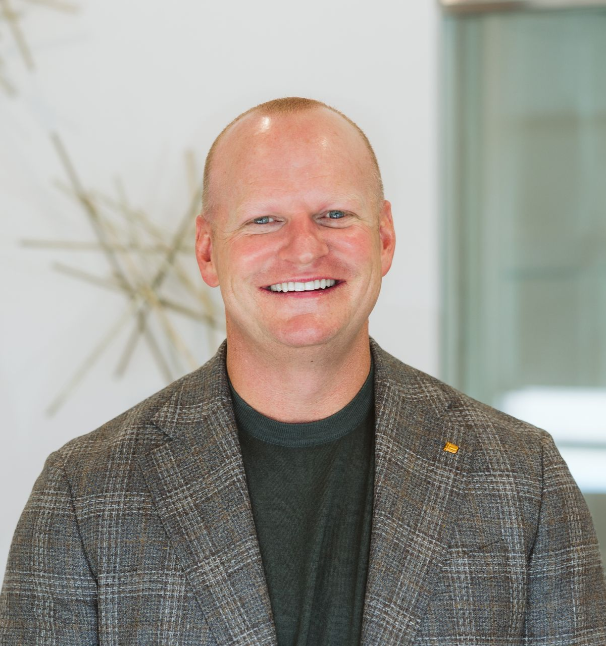 Smiling man in a gray plaid blazer standing indoors against a light background