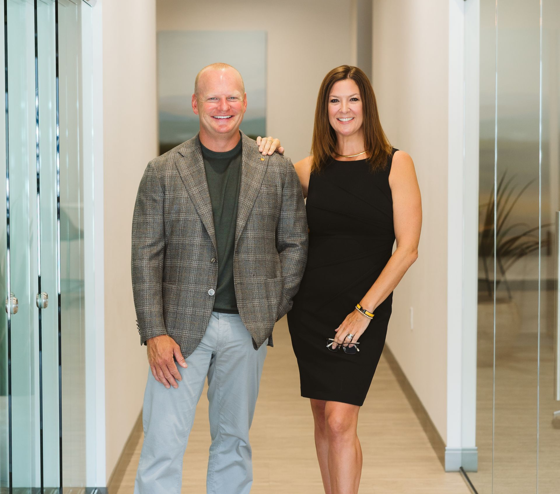Two people standing in a bright hallway, smiling side by side.