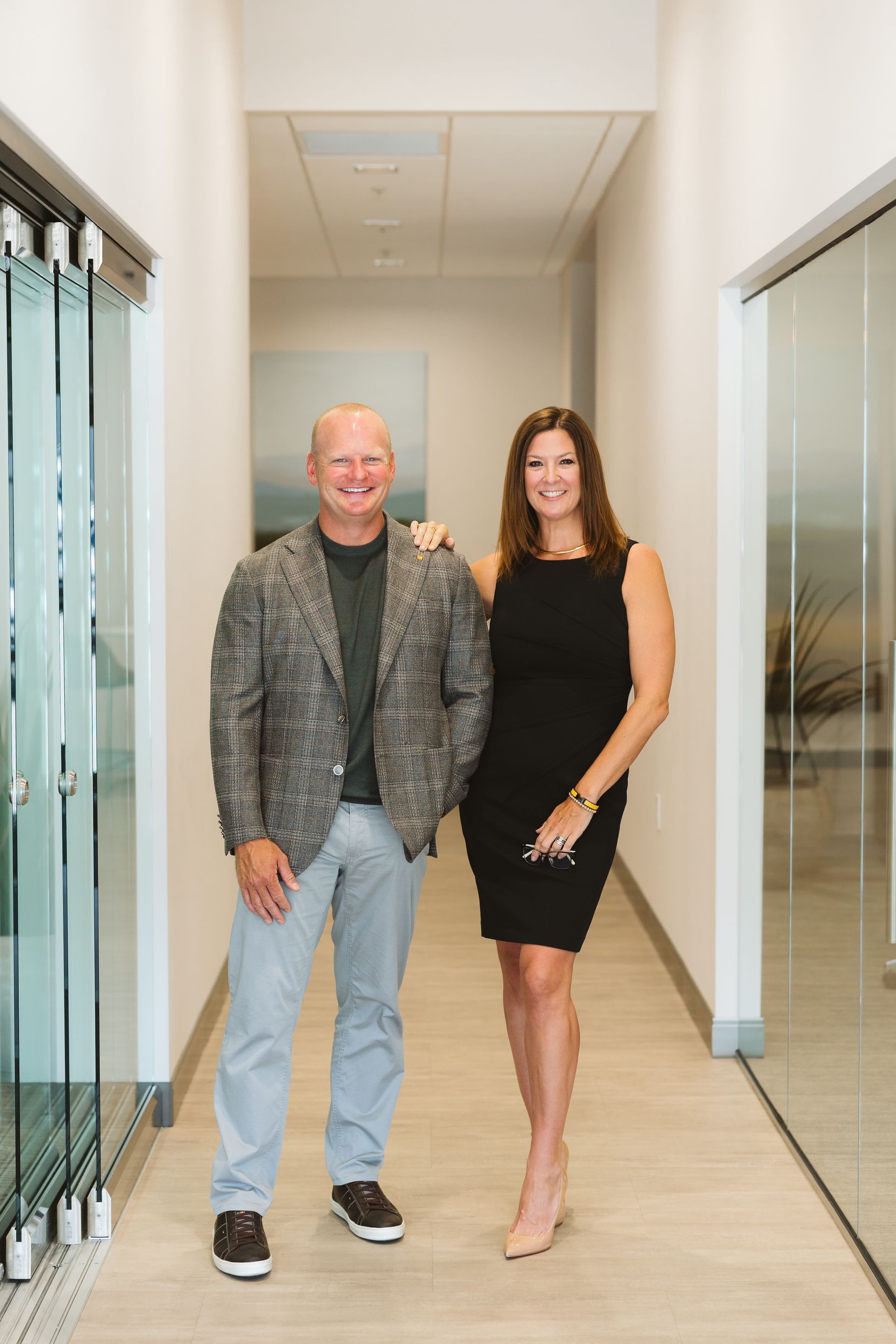 Two people standing in a bright office hallway, one in a gray jacket and jeans, the other in a black dress.