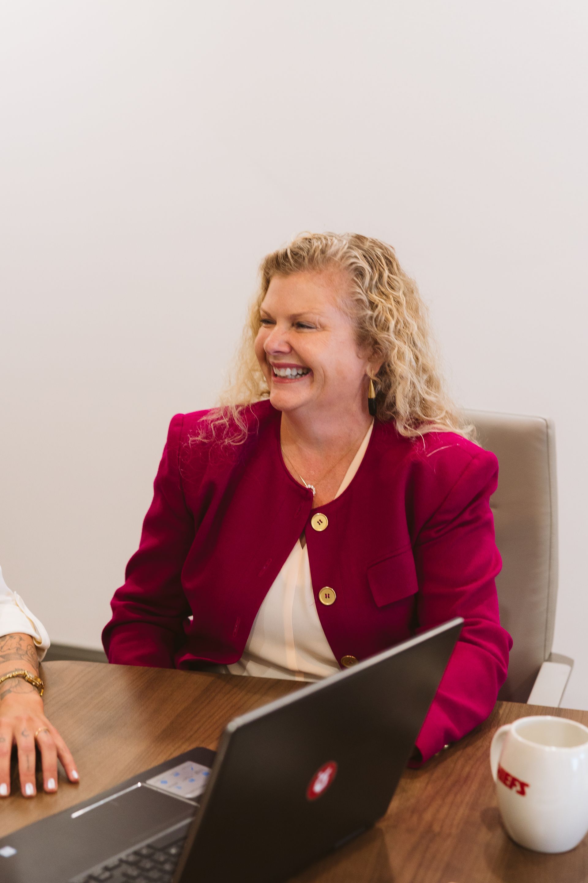 Woman in a red blazer smiling at a conference table with a laptop and coffee mug