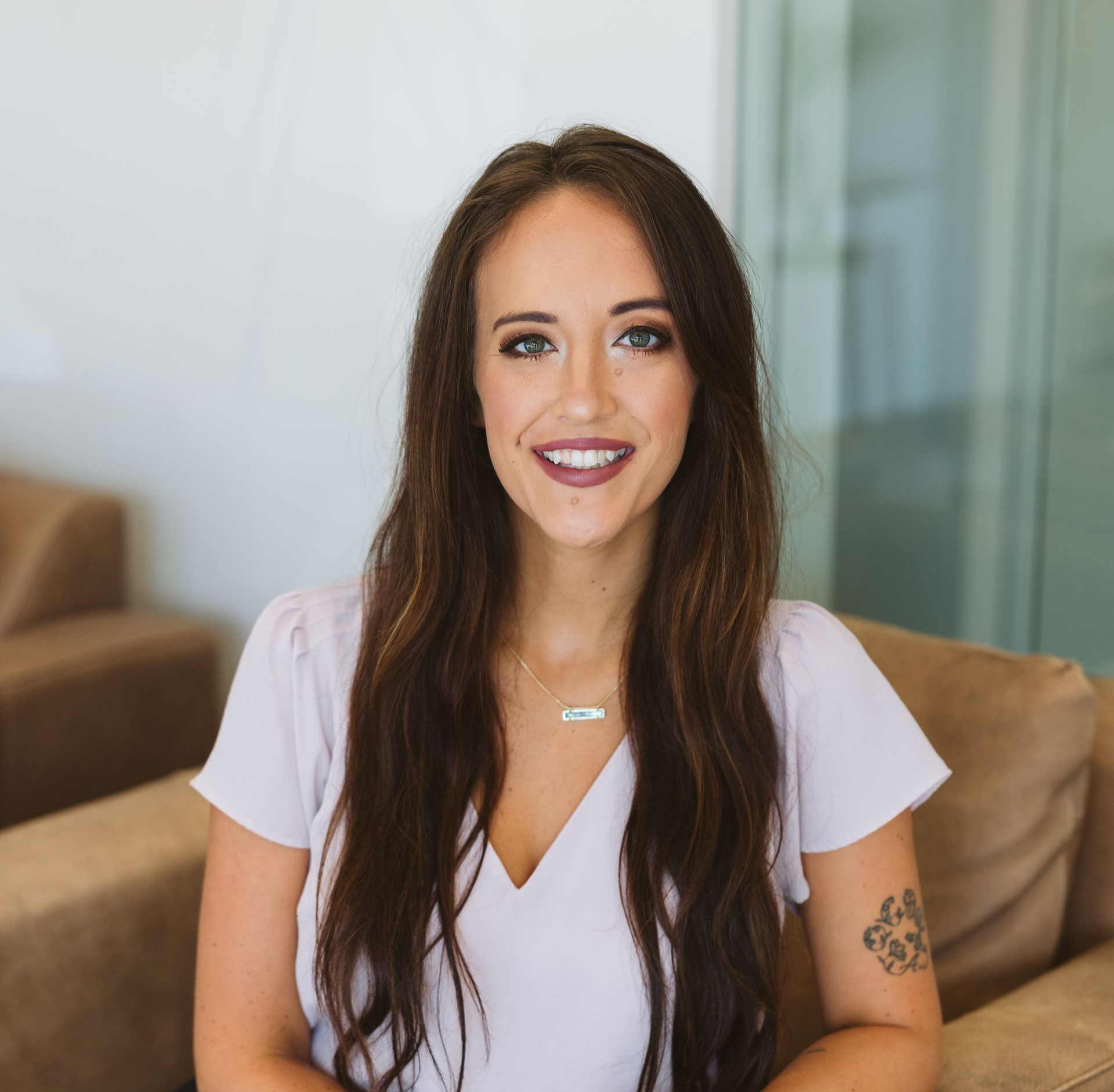 Woman smiling on a beige couch in a bright living room, wearing a white top.