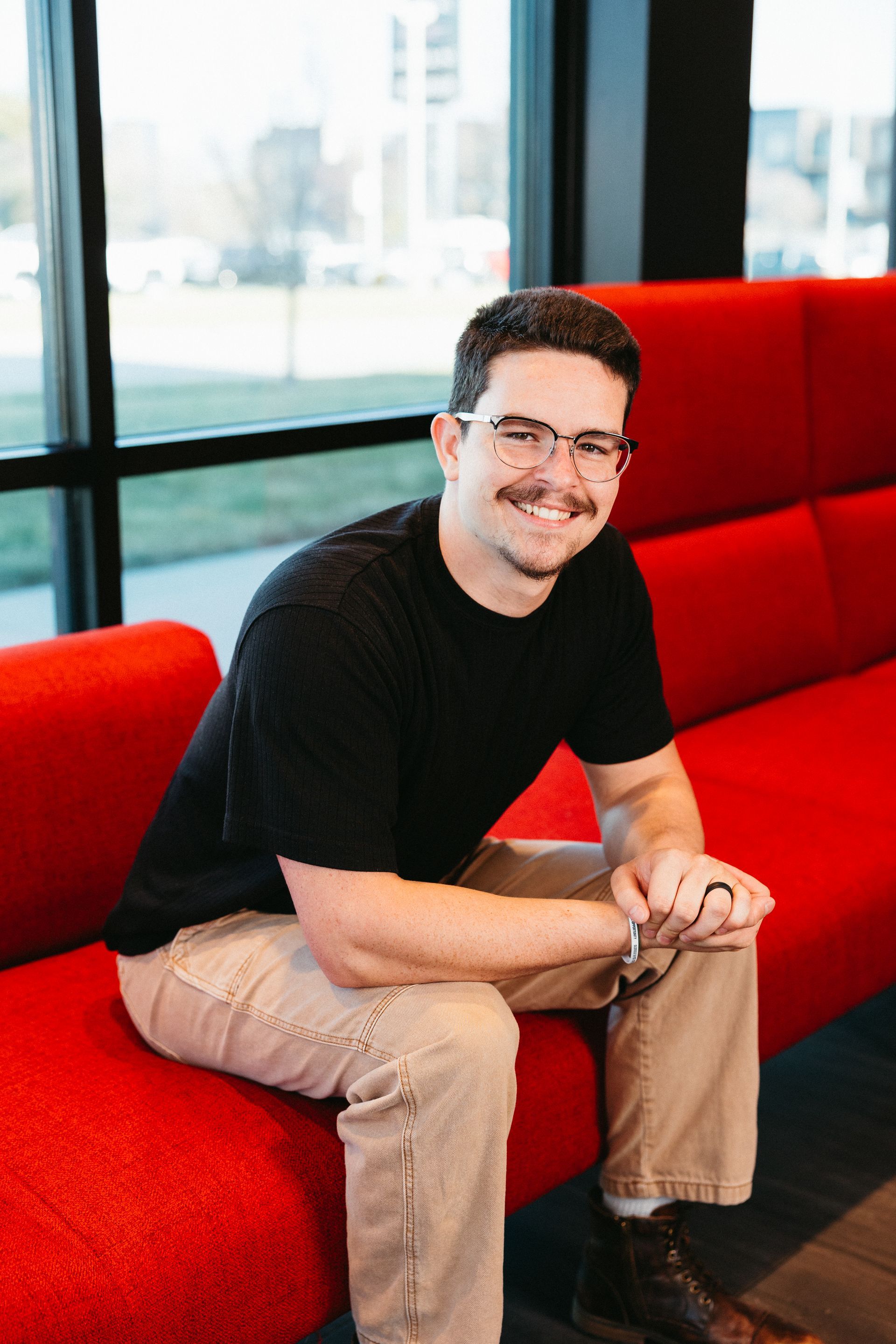 Smiling man in a black shirt sitting on a red couch by large windows.