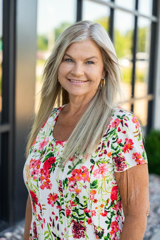 Smiling woman in a white floral blouse standing by a windowed wall outdoors