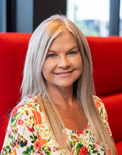 Smiling woman with long blonde hair in a floral top, seated before a red couch and window.