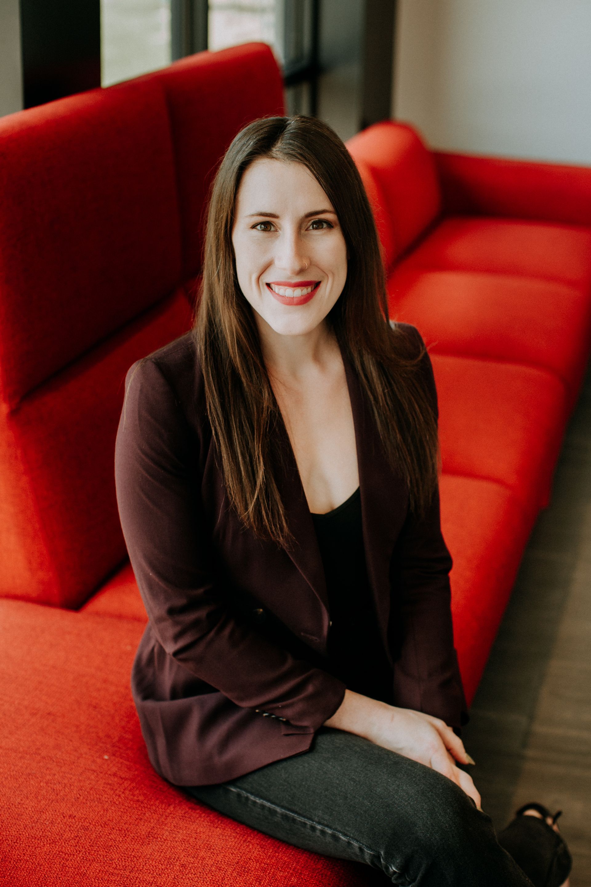 Woman in a burgundy blazer sitting on a red couch, smiling at the camera.