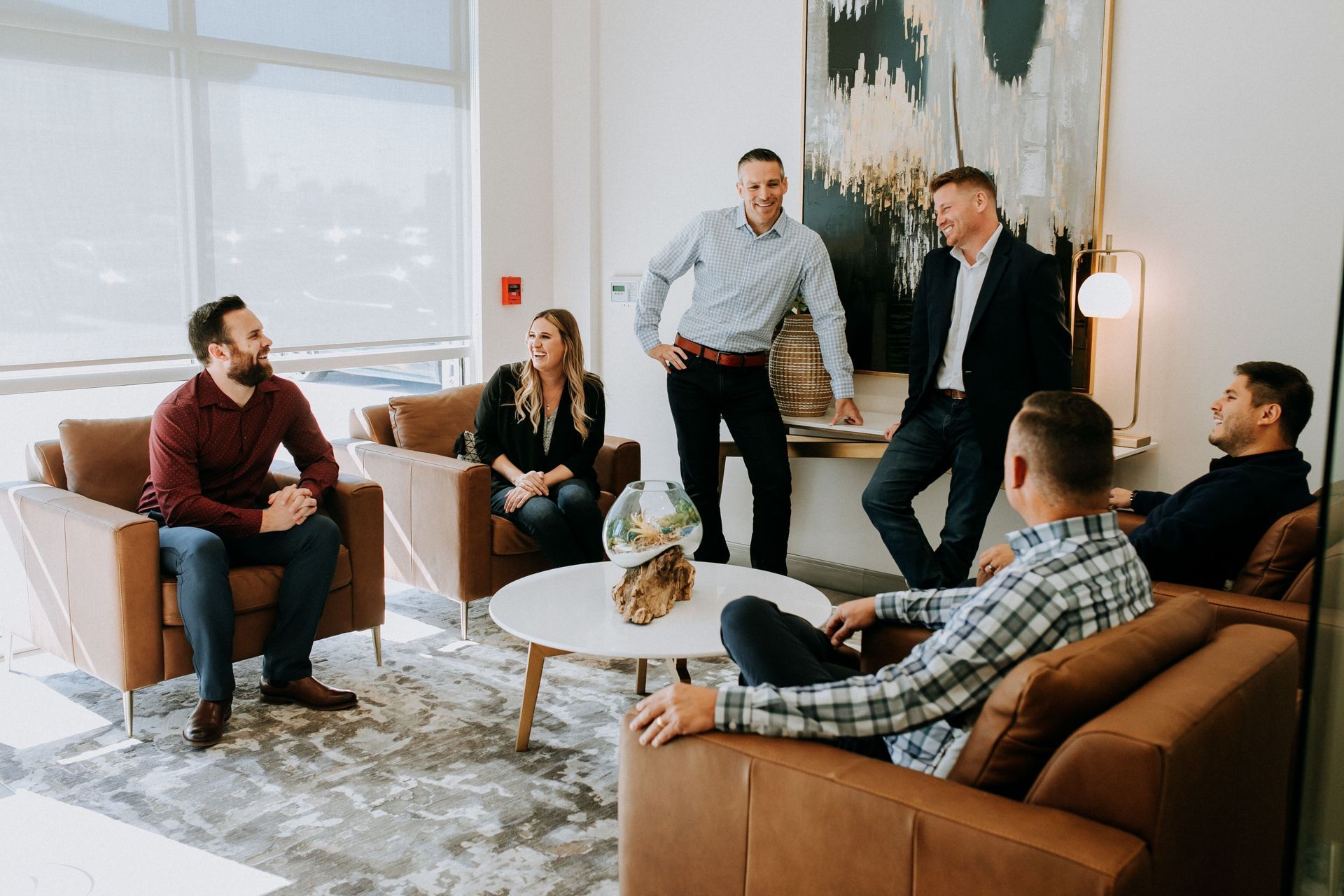 Five people chatting in a modern office lounge around brown chairs and a white coffee table.