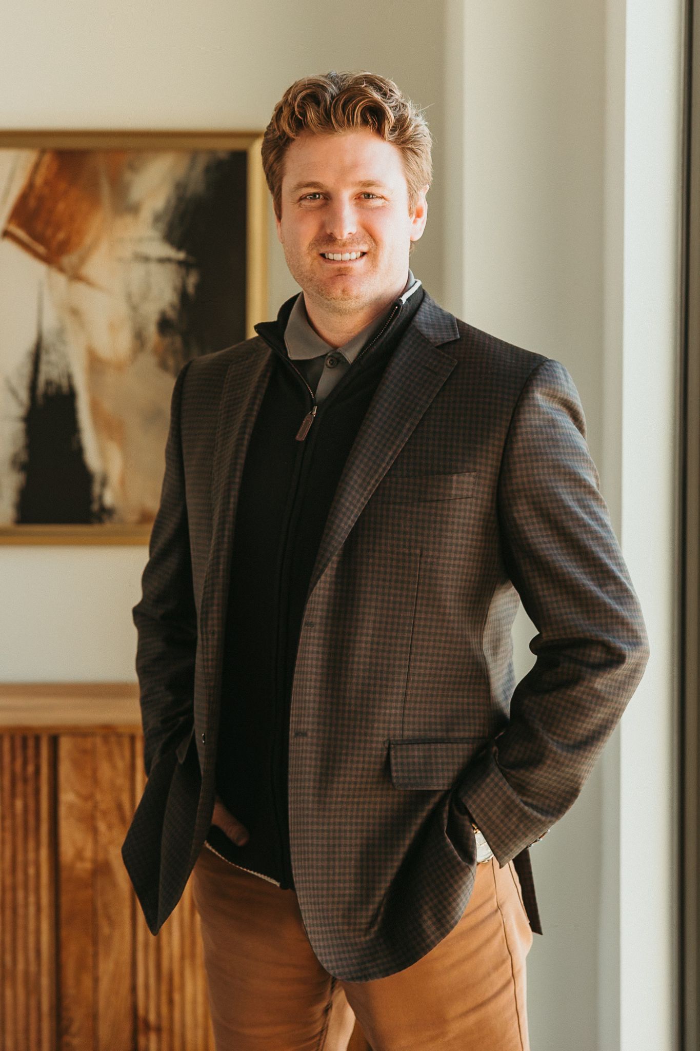 Smiling man in a dark blazer and brown pants stands indoors beside framed wall art.