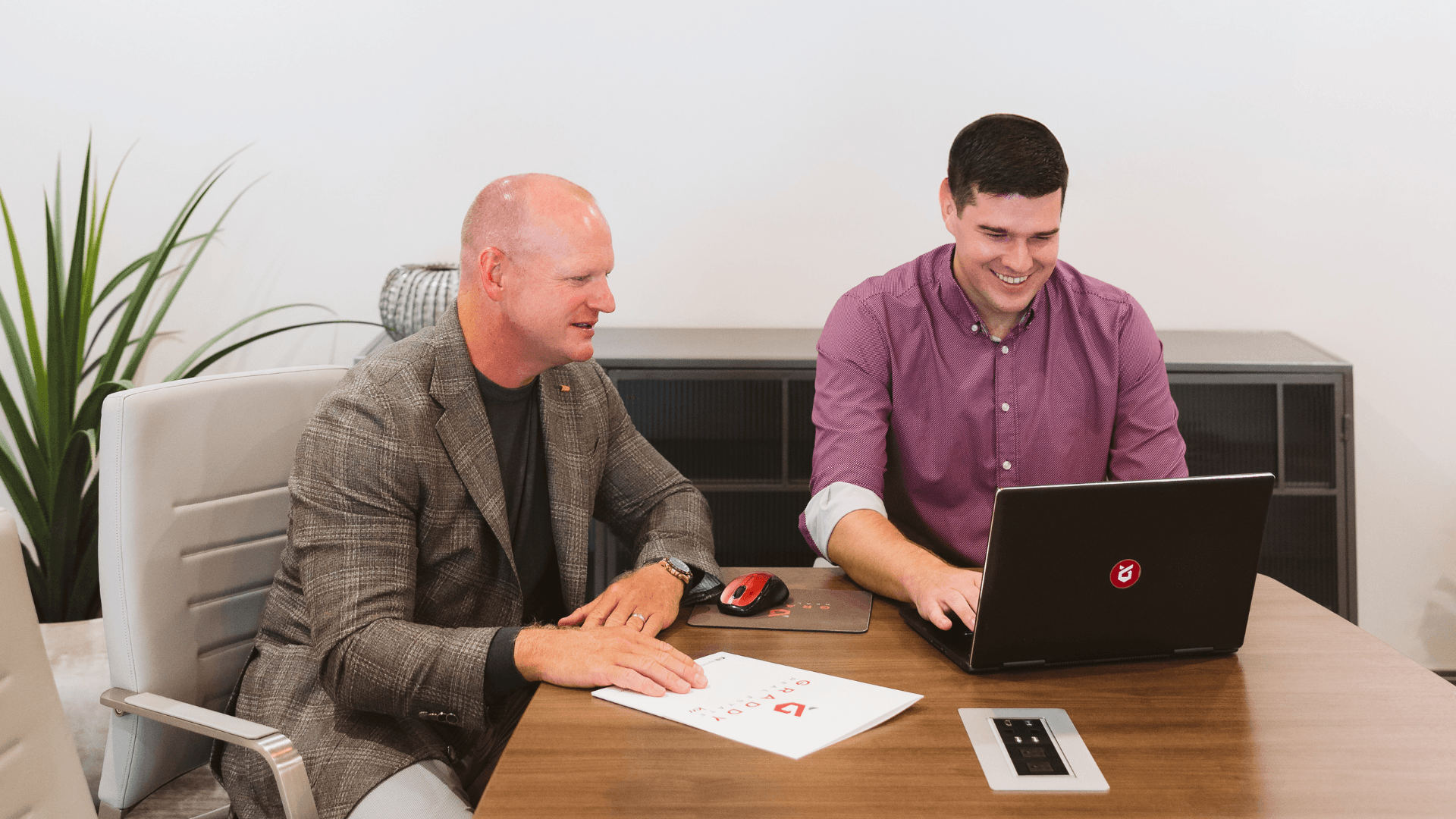 Two men at a desk reviewing papers and a laptop in a bright office