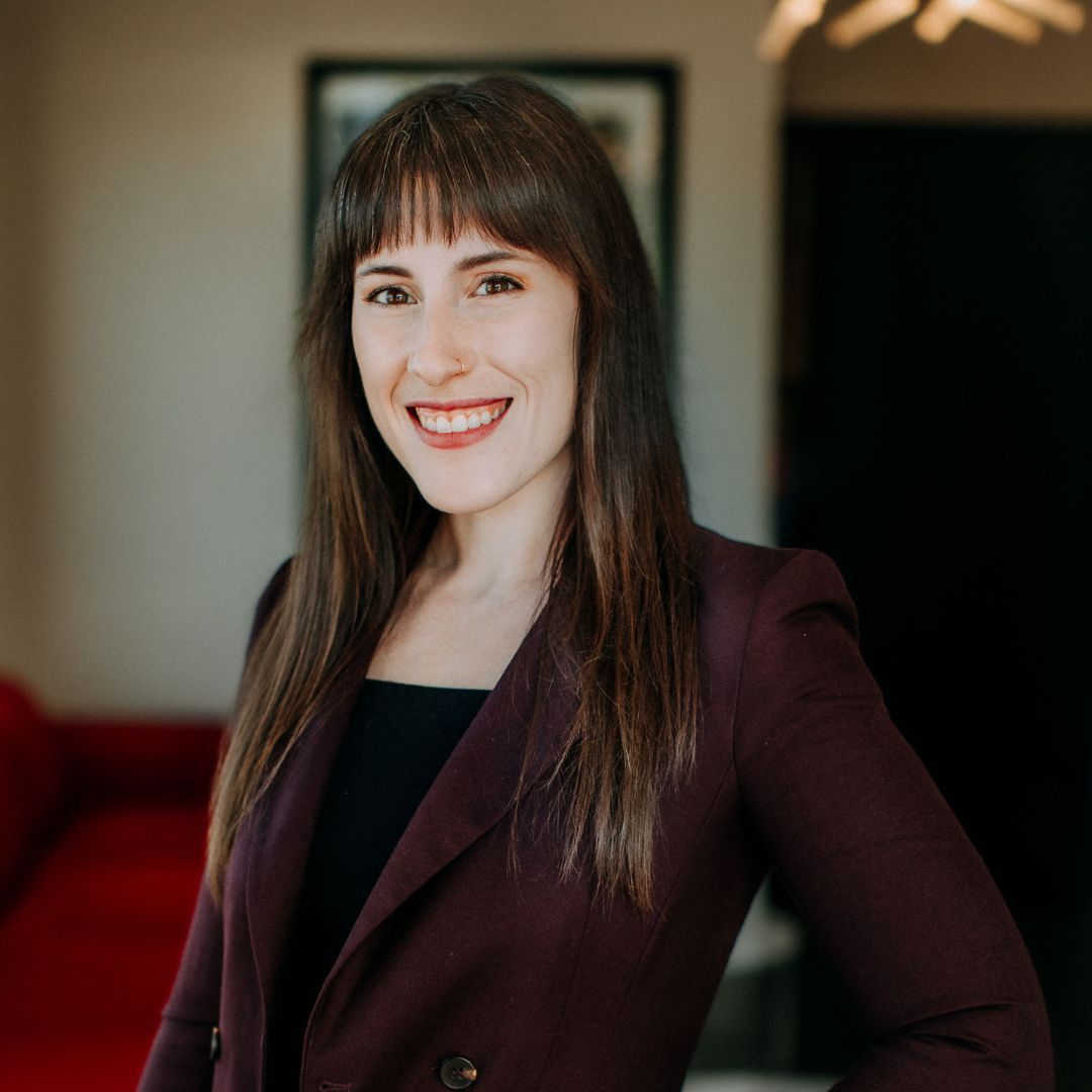 Smiling woman in a burgundy blazer standing indoors against a softly blurred background