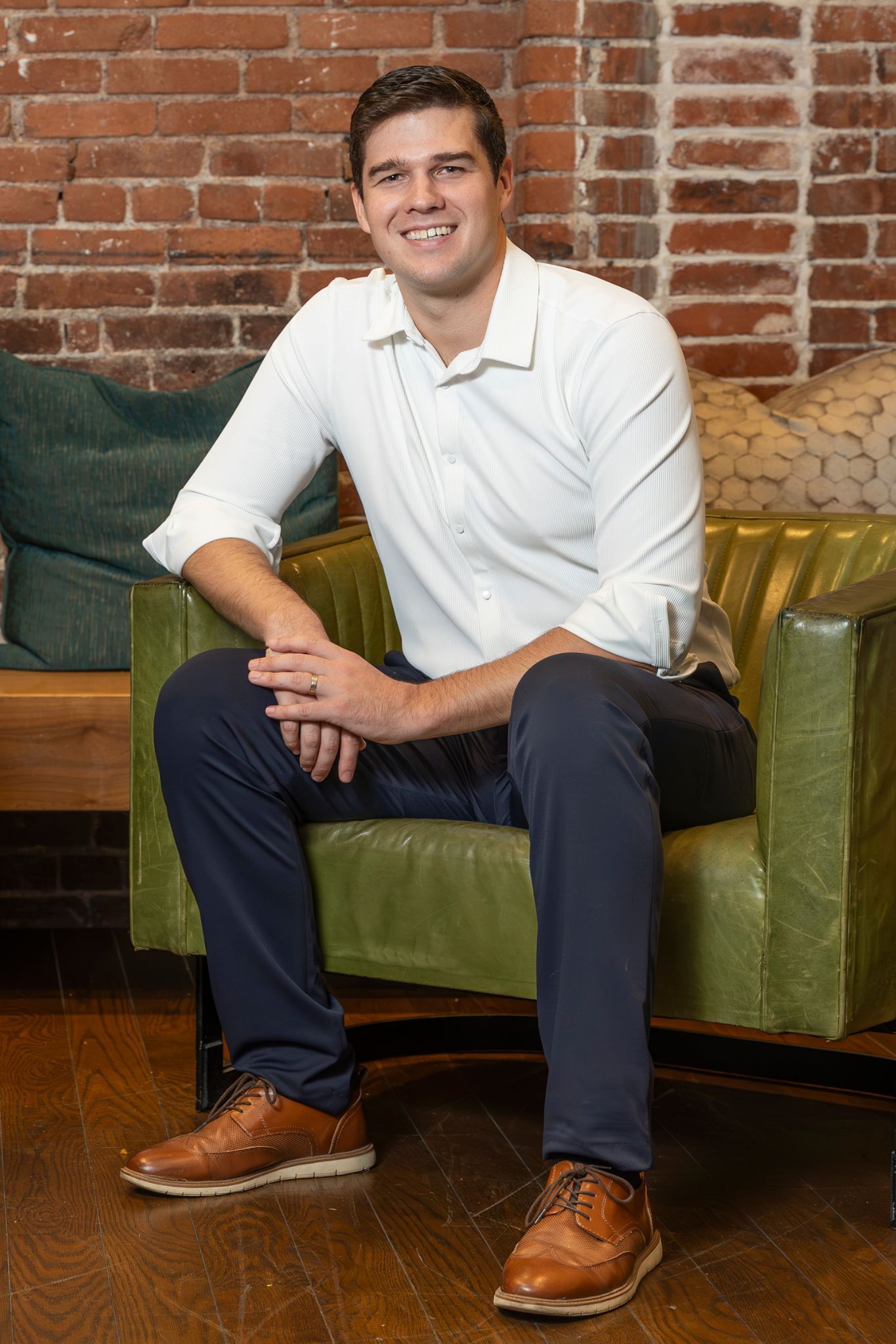 Smiling man sitting on a green couch against a brick wall, wearing a white shirt and dark pants.