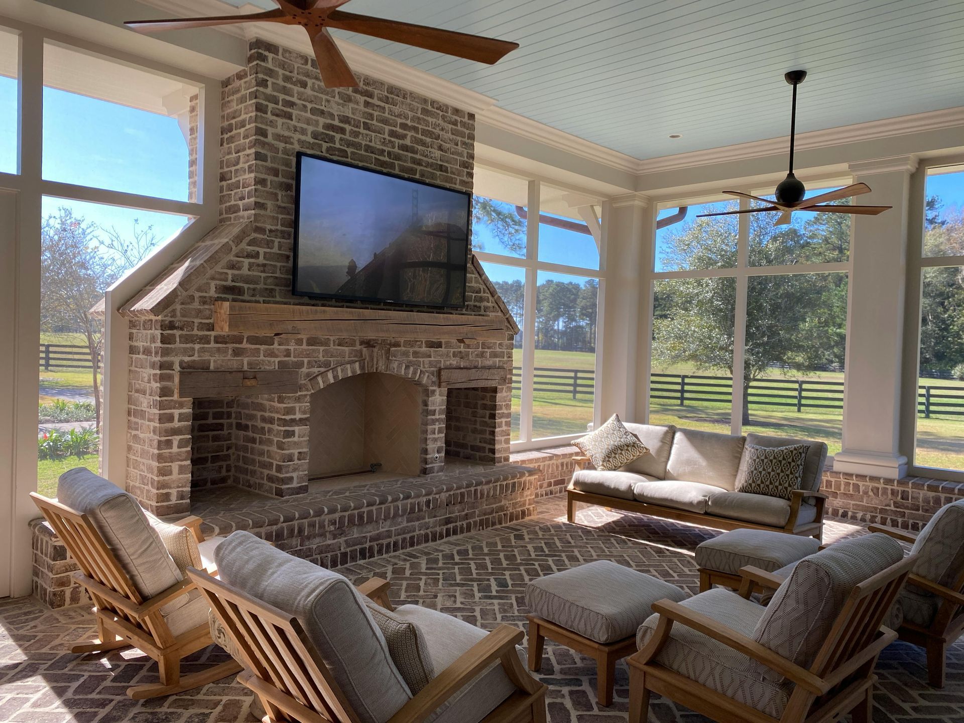 Sunlit brick patio room with TV over fireplace, wicker seating, and large windows overlooking a green yard