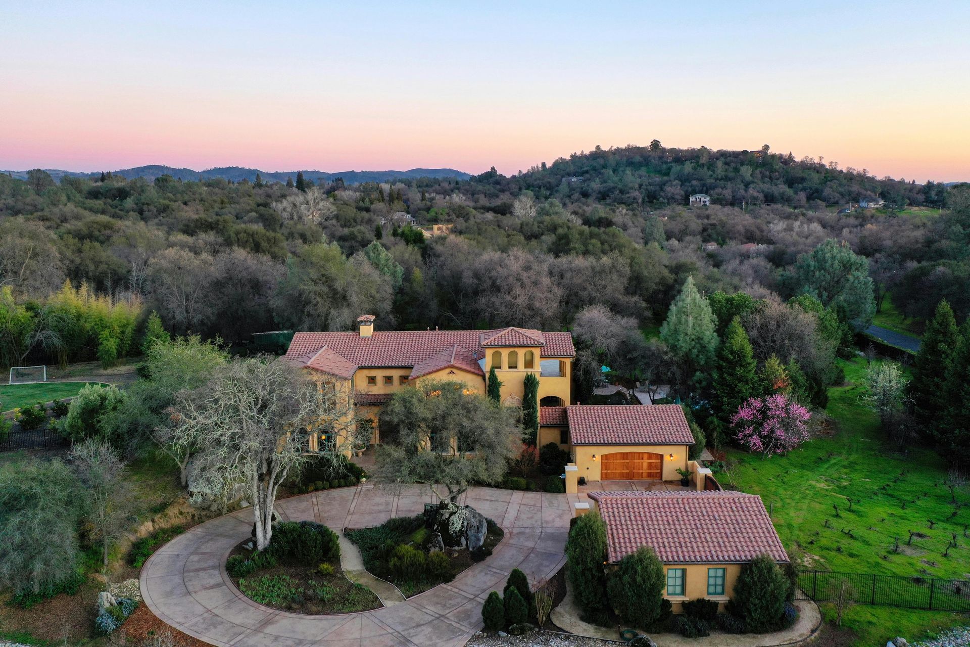 A rustic hillside villa with terracotta roofs, surrounded by trees and gardens at sunset.
